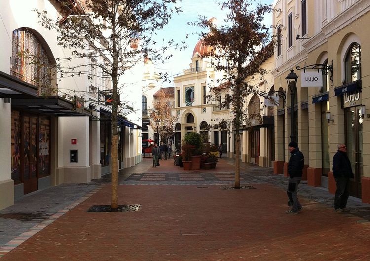 An outdoor shopping street with classic buildings, brick paving, and young trees.